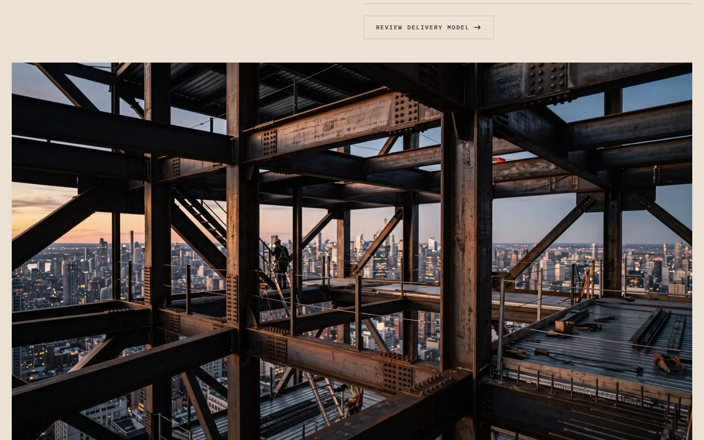 A photograph of a structural steel frame at height, the city skyline visible through it at dusk.